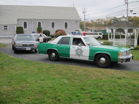 2001 Ford Crown Victoria - Multiple vehicles, including a police car, are parked in front of a building and gazebo.