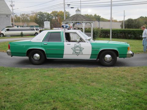 2001 Ford Crown Victoria - A two-tone green and white police car is parked on the side of the road, with a limousine visible in the background.