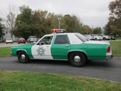2001 Ford Crown Victoria - A green and white sheriff's car, likely from the 1980s, is parked on a paved surface with other vehicles in the background.