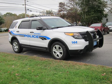 2001 Ford Crown Victoria - A white police SUV, marked "SWANSEA POLICE" with the unit number 144, is parked on a grassy area next to a road. Another car is visible in…