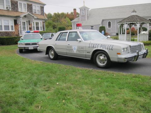 2001 Ford Crown Victoria - A gray Chrysler New Yorker police car, marked "Rhode Island State Police", is parked on a driveway next to a green police car.