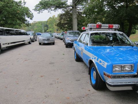 2001 Ford Crown Victoria - A line of vehicles including a police car and a limousine are parked on a street.