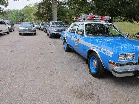2001 Ford Crown Victoria - A line of vehicles, including a vintage police car, are parked on an asphalt surface.