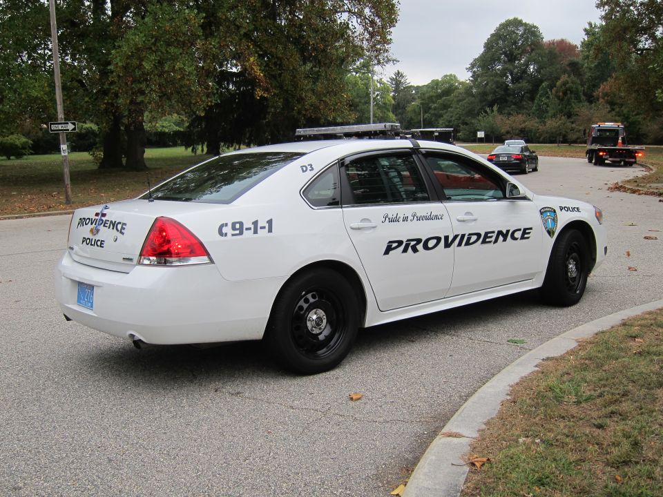 White police car with 'PROVIDENCE' and 'C9-1-1' markings. Another car and a tow truck are visible in the background