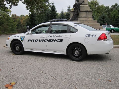 2001 Ford Crown Victoria - A white police car with "PROVIDENCE" on its side is parked near a monument, with another green vehicle in the background.