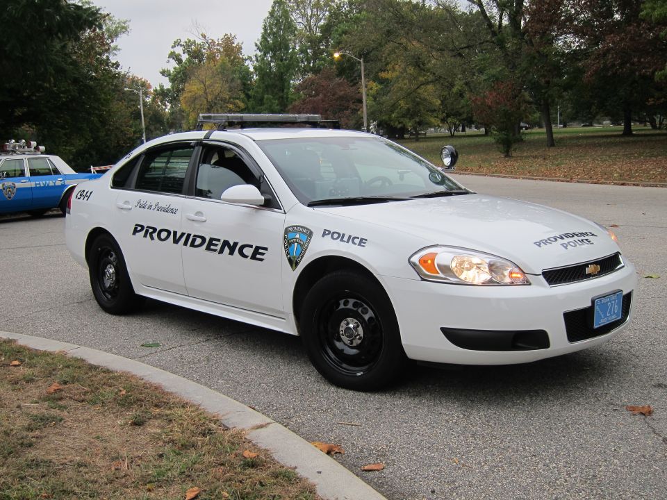 White police car with Providence Police markings and a blue license plate. Another blue and white police car is visible behind it