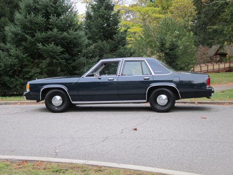 2001 Ford Crown Victoria - A dark blue sedan is parked on a street with trees and a bridge in the background.