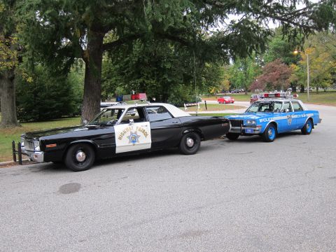 2001 Ford Crown Victoria - Two classic police cruisers, one black and one blue, are parked side-by-side on a paved road with trees in the background.