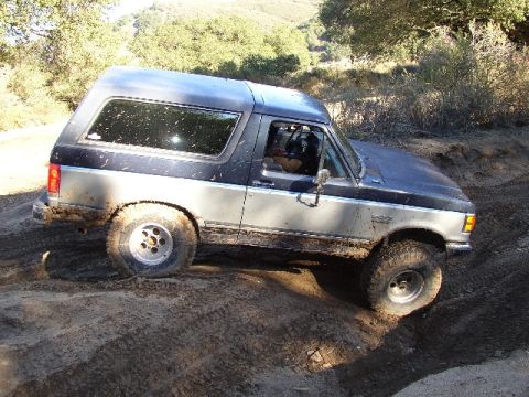 1989 Ford Bronco - Ford Bronco navigates a muddy off-road trail with trees in the background.
