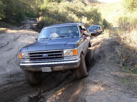 1989 Ford Bronco - A Ford F-Series truck navigates a challenging muddy off-road course with another vehicle following behind.