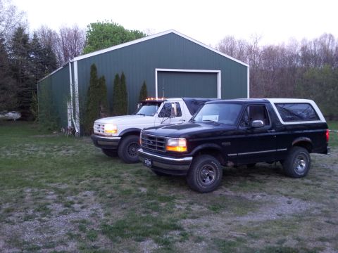 1995 Ford Bronco - Two Ford Broncos are parked in front of a dark green metal building with a white trim. The Bronco in the foreground is black with a white t…