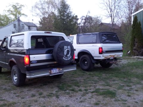 1995 Ford Bronco - Two Ford Broncos are parked in a grassy yard with houses and trees in the background. The Bronco on the left is dark colored with a white t…