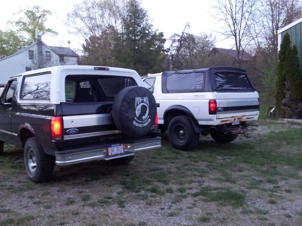 Two Ford Broncos parked in a yard, one dark, one white.