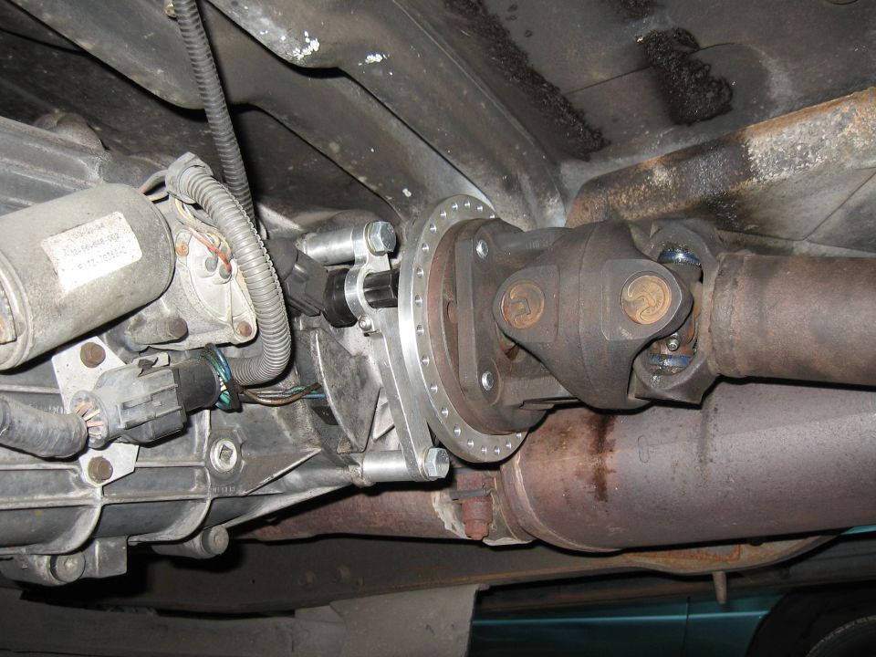 Underneath view of vehicle drivetrain: transfer case, driveshaft, adapter plate, exhaust.