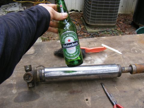 Hand holding Heineken bottle over a metal shaft with universal joint, wire brush, and pliers.