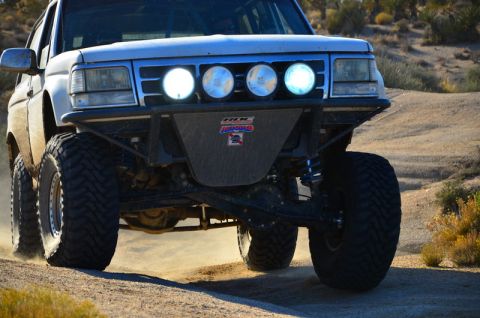 1996 Ford Bronco - The front quarter view of a white Ford Bronco modified for off-roading is kicking up dust on a dirt road. The vehicle features large, aggre…