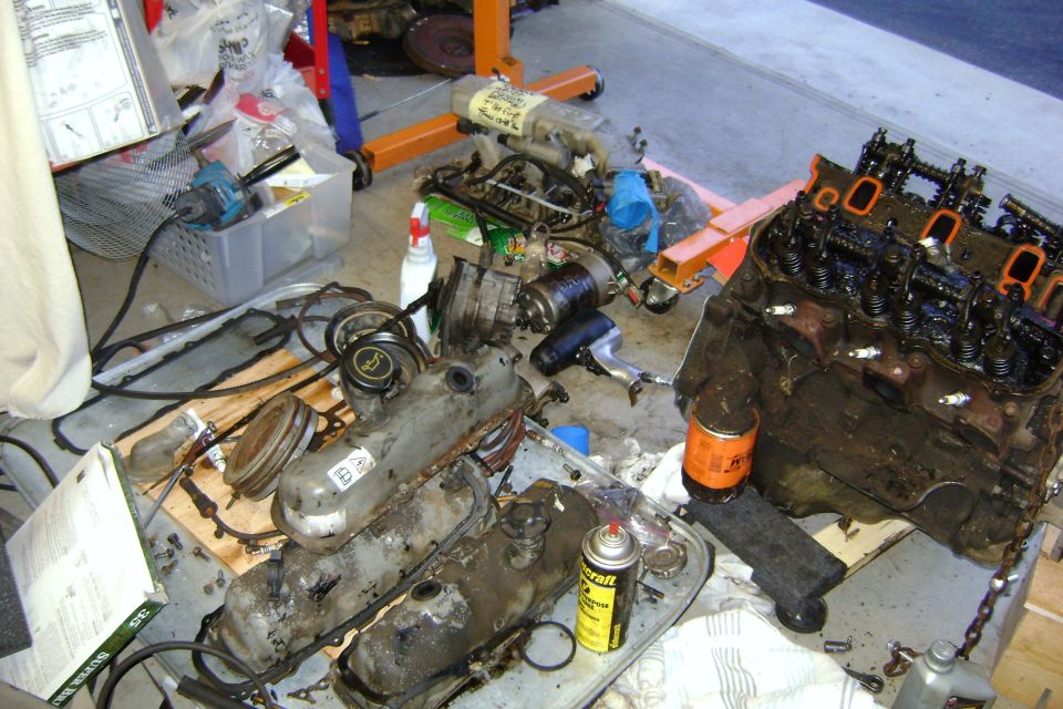 Engine block, valve covers, oil filter, and other engine parts scattered on a concrete floor with an engine hoist in the background