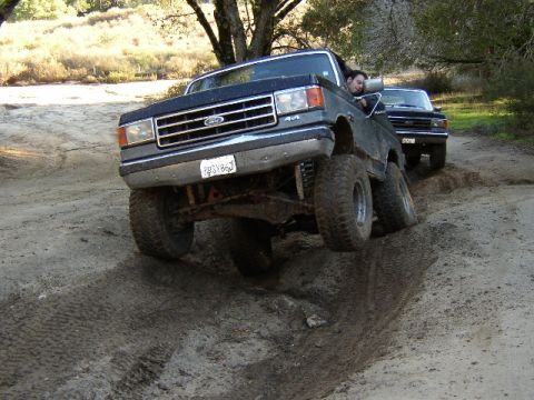 1989 Ford Bronco - Two Ford trucks navigate a muddy off-road trail with one vehicle at an extreme angle.