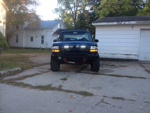 1996 Ford Bronco - The front view of a dark blue Ford Bronco is parked in a driveway in front of a white garage. The Bronco has a custom black bumper with aux…