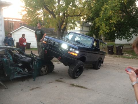 1996 Ford Bronco - A lifted, dark blue Ford Bronco is parked on top of a heavily damaged green car. Three men stand nearby, observing the scene.