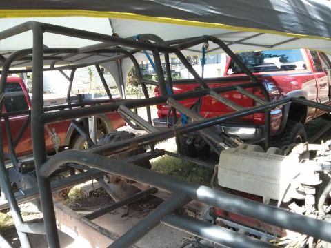 1986 Ford Bronco - A custom roll cage is being built around the chassis of a vehicle, with a red pickup truck visible in the background under a yellow and gra…