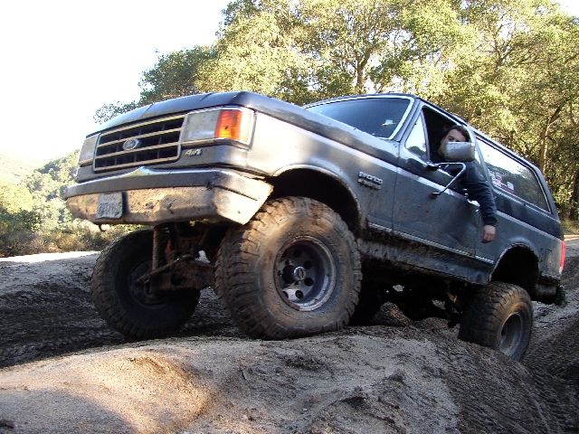 Gray lifted Ford Bronco on muddy tires climbing a dirt mound off-road, front-quarter view