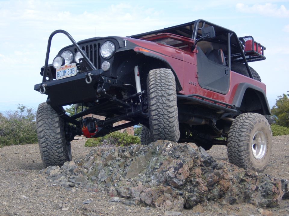 Red Jeep CJ7 with custom bumper and roll cage driving over rocks.