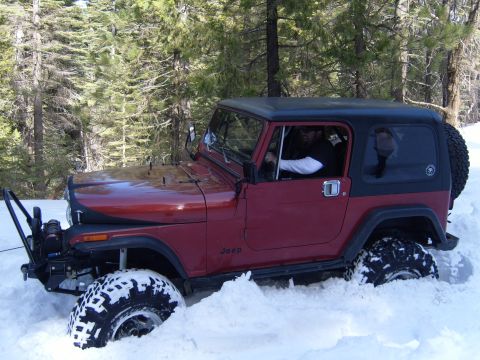 1985 Jeep CJ7 - A red Jeep CJ7 is stuck in deep snow with its front tire partially buried. The driver is visible behind the steering wheel, wearing sunglas…