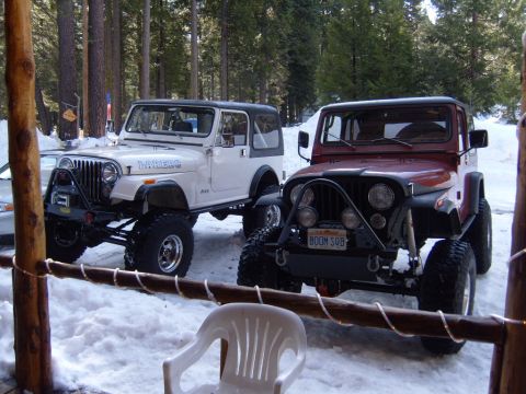 1985 Jeep CJ7 - Two modified Jeep CJ-style vehicles are parked in the snow, surrounded by pine trees. The Jeep on the left is white with a black top and "L…