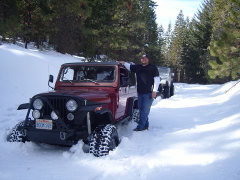 1985 Jeep CJ7 - A man stands next to a red Jeep CJ7 partially submerged in snow, with a white Jeep visible behind it on a snow-covered road surrounded by p…