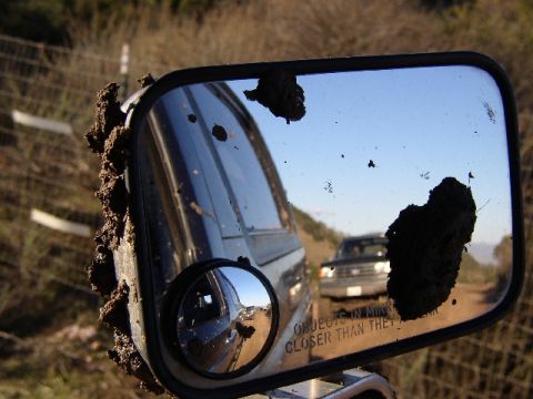 1989 Ford Bronco - Side mirror caked with mud, reflecting another vehicle on a dusty trail.