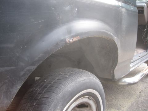 1993 Ford Bronco - Close-up side view of the rear wheel well area of a dark colored vehicle, showing rust on the fender flare and a chrome step bar below.