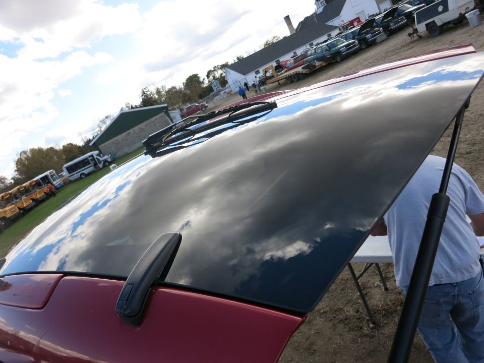 Car hood reflecting clouds, person in blue shirt behind it. Other vehicles and buses in background