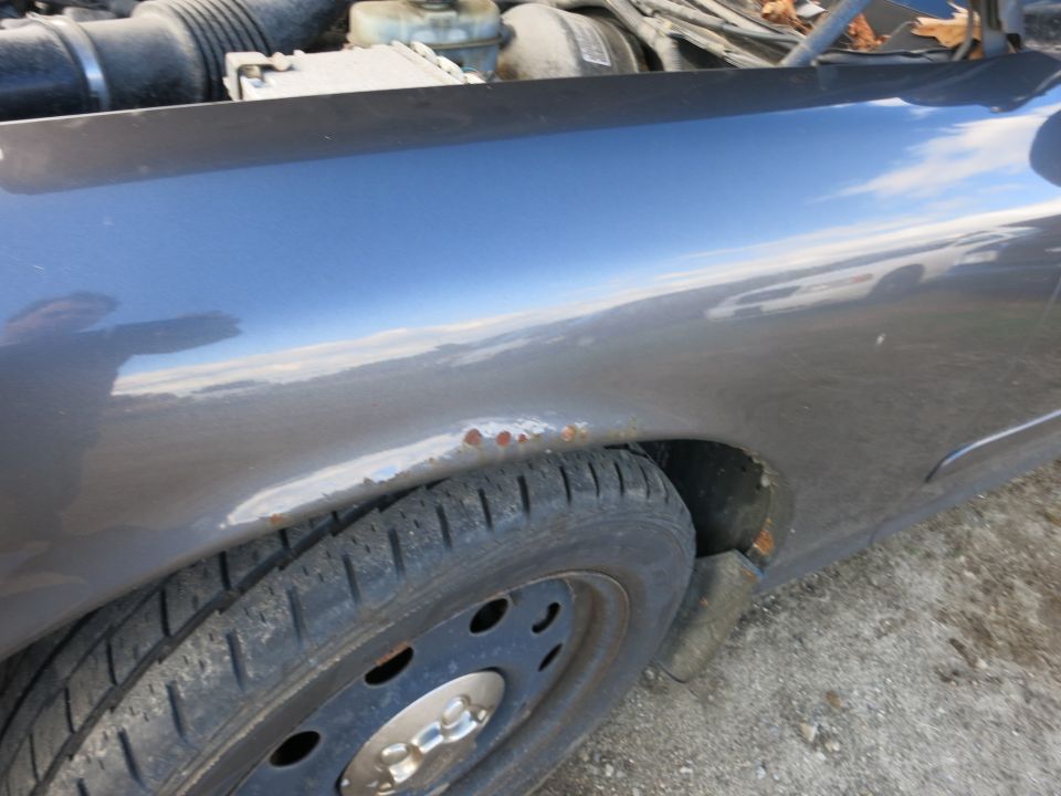 Car fender showing rust spots near the wheel well. Engine components are visible under the hood