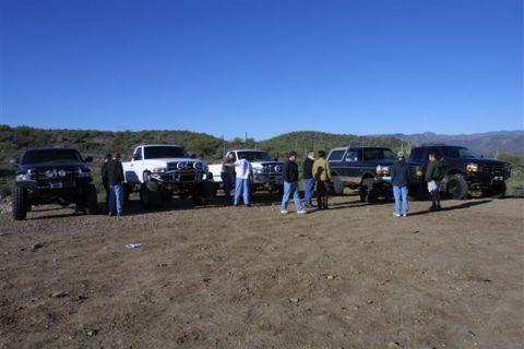 1994 Ford Bronco - A group of modified trucks and SUVs are gathered in a desert area under a clear blue sky.