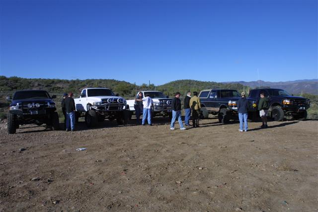 Four lifted trucks and Broncos parked in a row on desert dirt with people standing between them