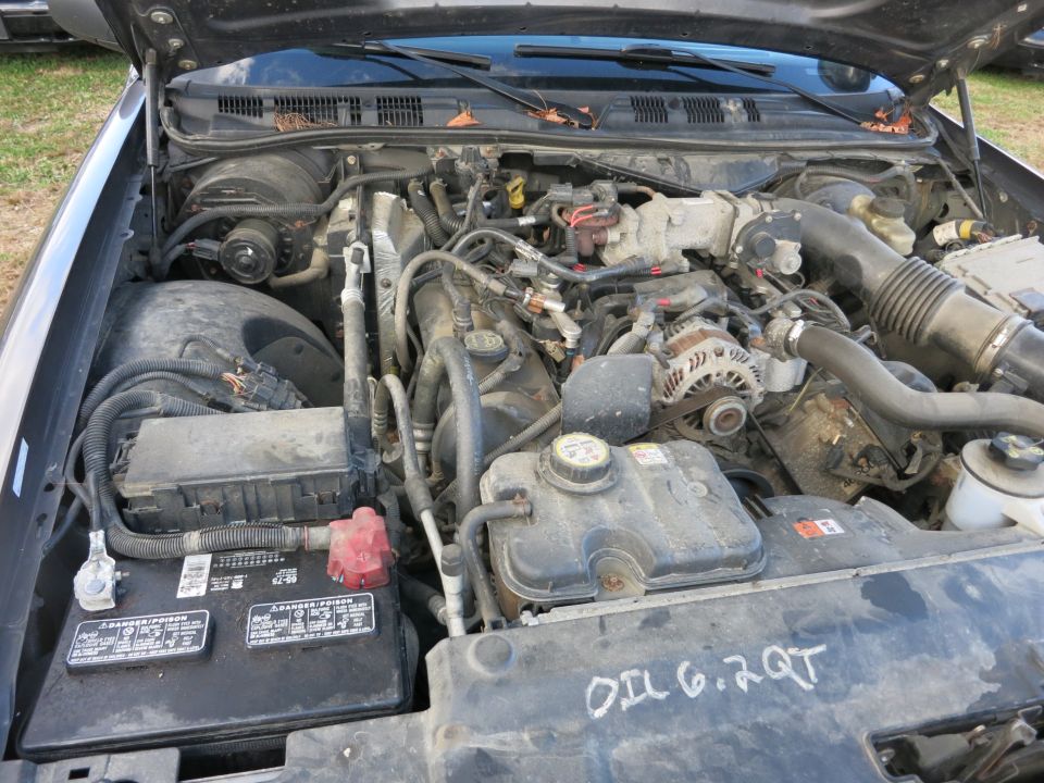 Engine bay of a Ford Crown Victoria, showing the V8 engine, battery, and coolant reservoir. Text on the cover reads 'OIL 6.2 QT'