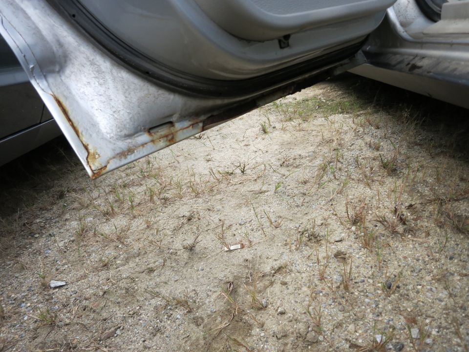 Opened car door showing rusted sill and interior floor with sand and grass