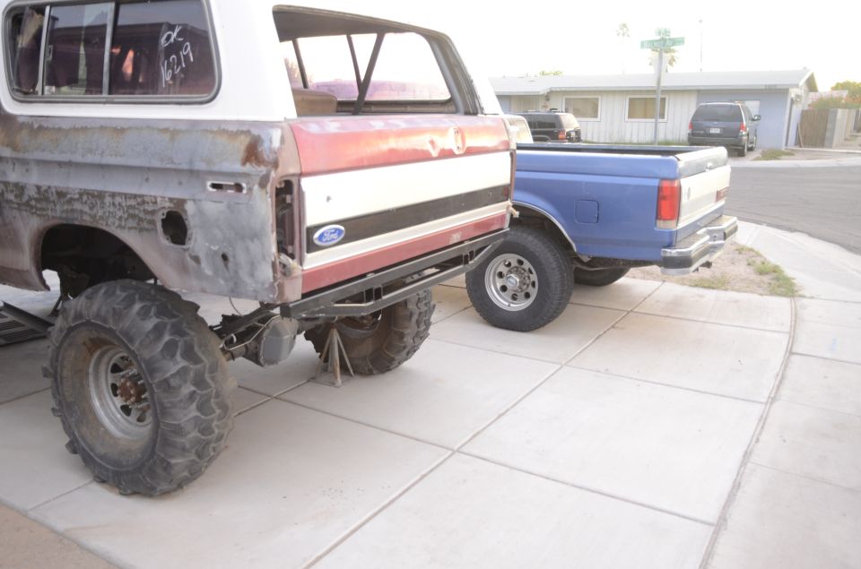 Rear view of a modified Ford Bronco with large tires, next to a blue Ford pickup truck.