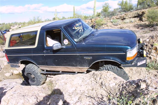 Dark green 1990s Ford Bronco with tan top flexing over desert rocks among saguaro cactus