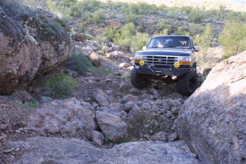 1994 Ford Bronco - A dark blue Ford Bronco is shown off-roading on a rocky, desert-like terrain.