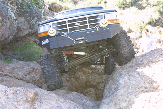 Modified dark Ford 4x4 with red coil-overs crawling over boulders, front three-quarter.