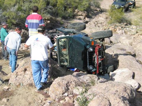 1994 Ford Bronco - An off-road vehicle has overturned in a rocky, desert-like environment, with onlookers assessing the situation.