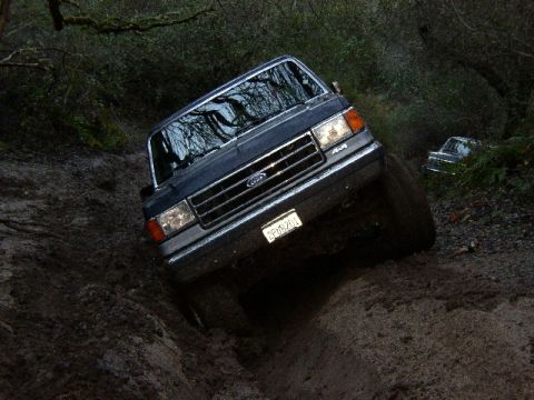 1989 Ford Bronco - A dark blue Ford F-Series truck navigates a muddy off-road trail, with another vehicle visible in the background.