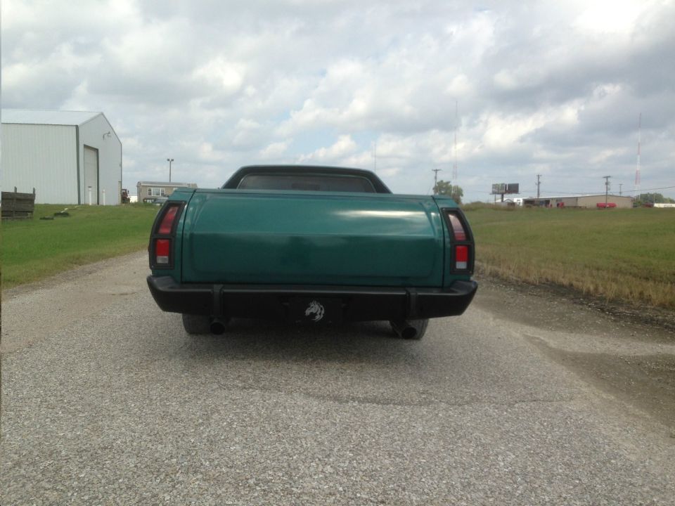 Green pickup truck rear view, black bumper with horse emblem, dual exhaust pipes, gravel road