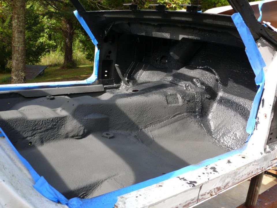 Car interior floor pan coated in gray primer, with blue painter's tape along the edges