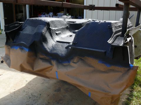 Car chassis part covered in black spray paint and masking paper, suspended by metal frame